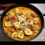 A close-up of rich, creamy One-Pot Sausage Tortellini Harvest Soup topped with grated cheese in a dark pot.