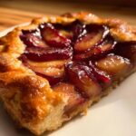 A close-up, golden-lit slice of Plum Rustic Tart showing glossy, caramelized plum slices on a flaky crust.
