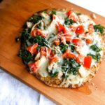 A finished Quick Chicken Flatbread Dinner topped with melted cheese, diced tomatoes, and kale on a wooden board.