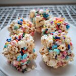 Four colorful Rainbow Marshmallow Popcorn Balls studded with bright sprinkles, resting on a white plate.
