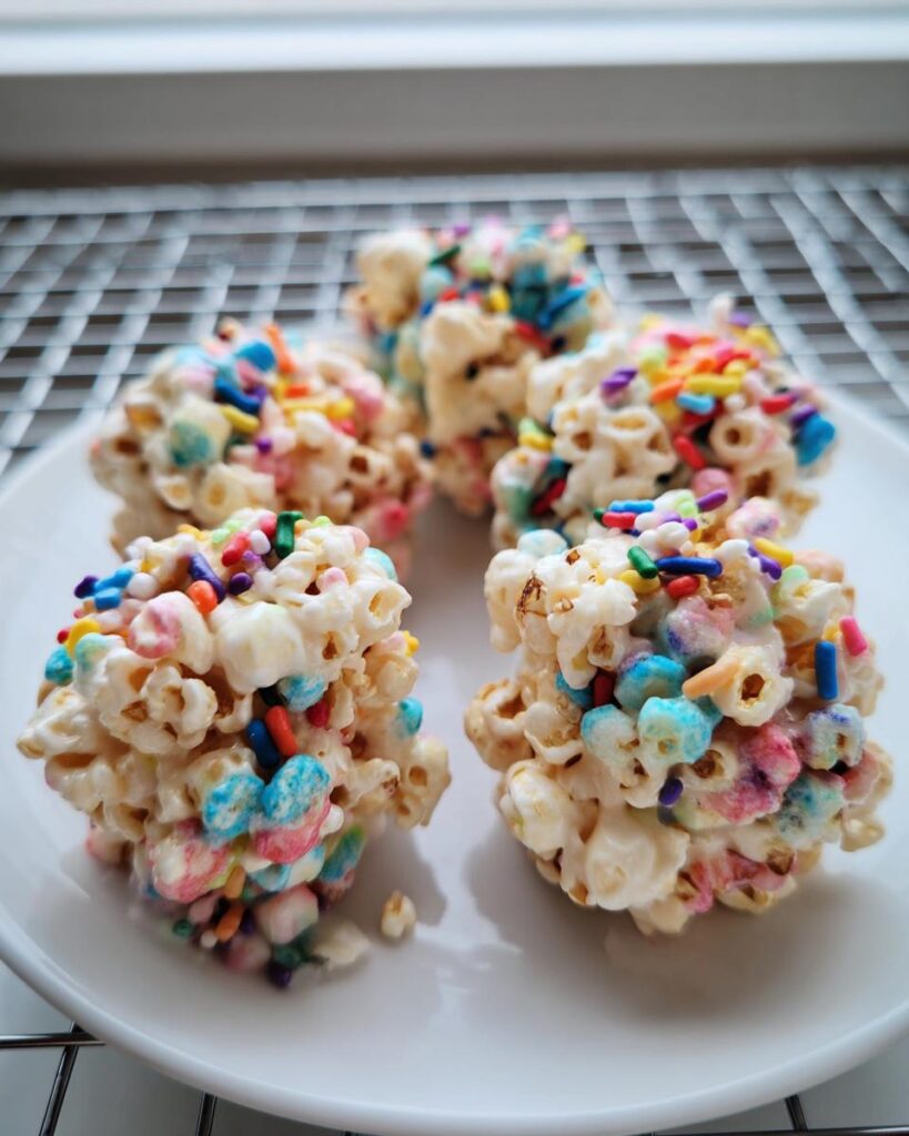 Four colorful Rainbow Marshmallow Popcorn Balls topped with sprinkles, resting on a white plate.