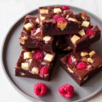 A stack of fudgy Raspberry White Chocolate Brownies topped with white chocolate chunks and fresh raspberries.