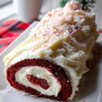 Cross-section view of a Red Velvet Christmas Log Cake, showing the red sponge rolled with white cream cheese frosting and topped with crushed peppermint.