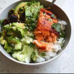 A close-up of a finished Salmon Avocado Protein Bowl featuring flaky salmon over rice, topped with avocado, Brussels sprouts, and sesame seeds.