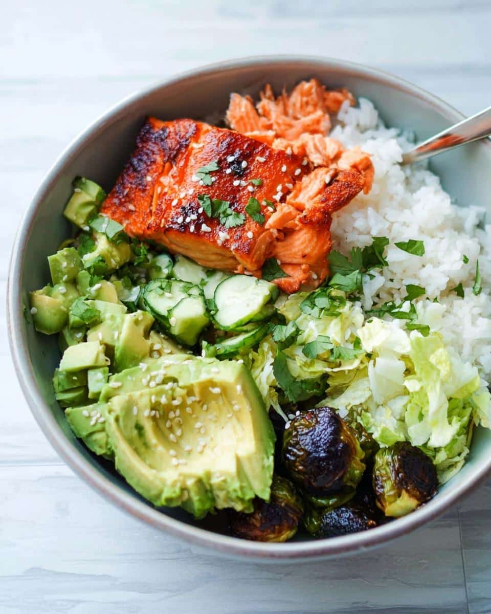 Close-up of a Salmon Avocado Protein Bowl featuring a piece of glazed salmon, sliced avocado, white rice, cucumbers, and roasted Brussels sprouts.
