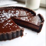 A rich, dark Chocolate Ganache Tart topped with flaky sea salt, with one slice cut and slightly pulled away.