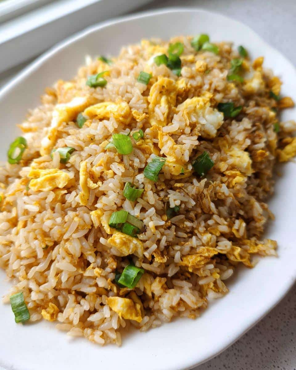 A close-up shot of a white plate piled high with Simple Egg Fried Rice, featuring scrambled egg pieces and topped with fresh green scallions.