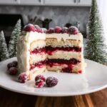 A slice of layered CRANBERRY WHITE CHRISTMAS CAKE with white frosting and sugared cranberries, set next to miniature Christmas trees.