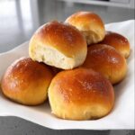 A close-up of several golden brown, shiny Soft Thermomix Dinner Rolls stacked on a white napkin.