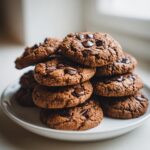 A tempting stack of freshly baked, dark brown Thermomix Chocolate Chip Cookies piled high on a white plate.