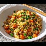 A close-up of a white bowl filled with healthy Cauliflower Fried Rice, mixed with scrambled egg, bright green peas, and diced carrots.