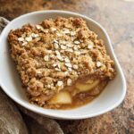 A square slice of Thermomix Apple Crumble Cake with a thick, crumbly oat topping served in a white bowl.
