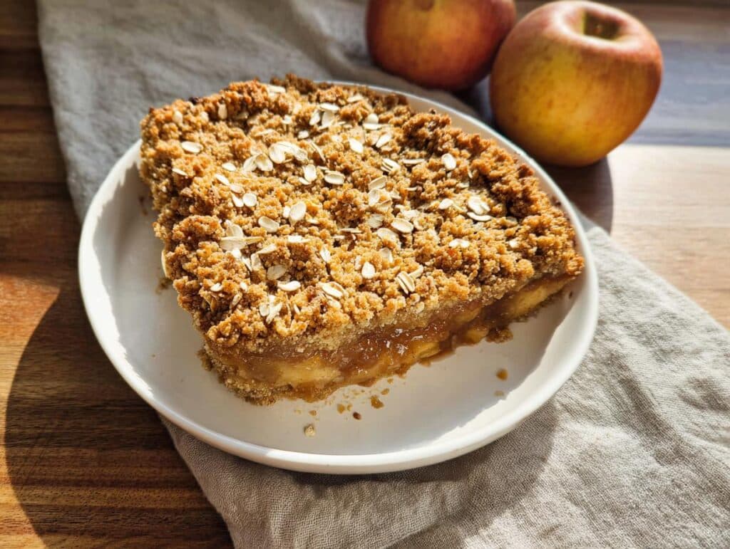 A close-up of a slice of Thermomix Apple Crumble Cake on a white plate, topped with oat crumble.
