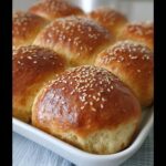 A close-up of golden brown Thermomix Brioche Buns topped with sesame seeds, arranged in a white baking dish.