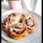 Five freshly baked Thermomix Cinnamon Scrolls topped with white icing and chopped walnuts, served on a white plate.