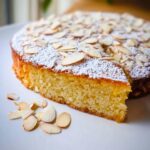 A slice cut from the TM6 Orange & Almond Flourless Cake, dusted with powdered sugar and topped with flaked almonds.