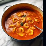 A close-up of rich Tomato Tortellini Soup simmering in a blue enameled Dutch oven, garnished with herbs.