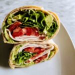 A cross-section view of a Turkey & Avocado Whole Wheat Wrap cut in half, showing layers of turkey, tomato, lettuce, and avocado spread.