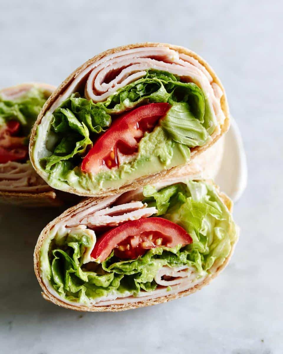 Close-up of a Turkey & Avocado Whole Wheat Wrap cut in half, showing layers of turkey, lettuce, tomato, and avocado spread.