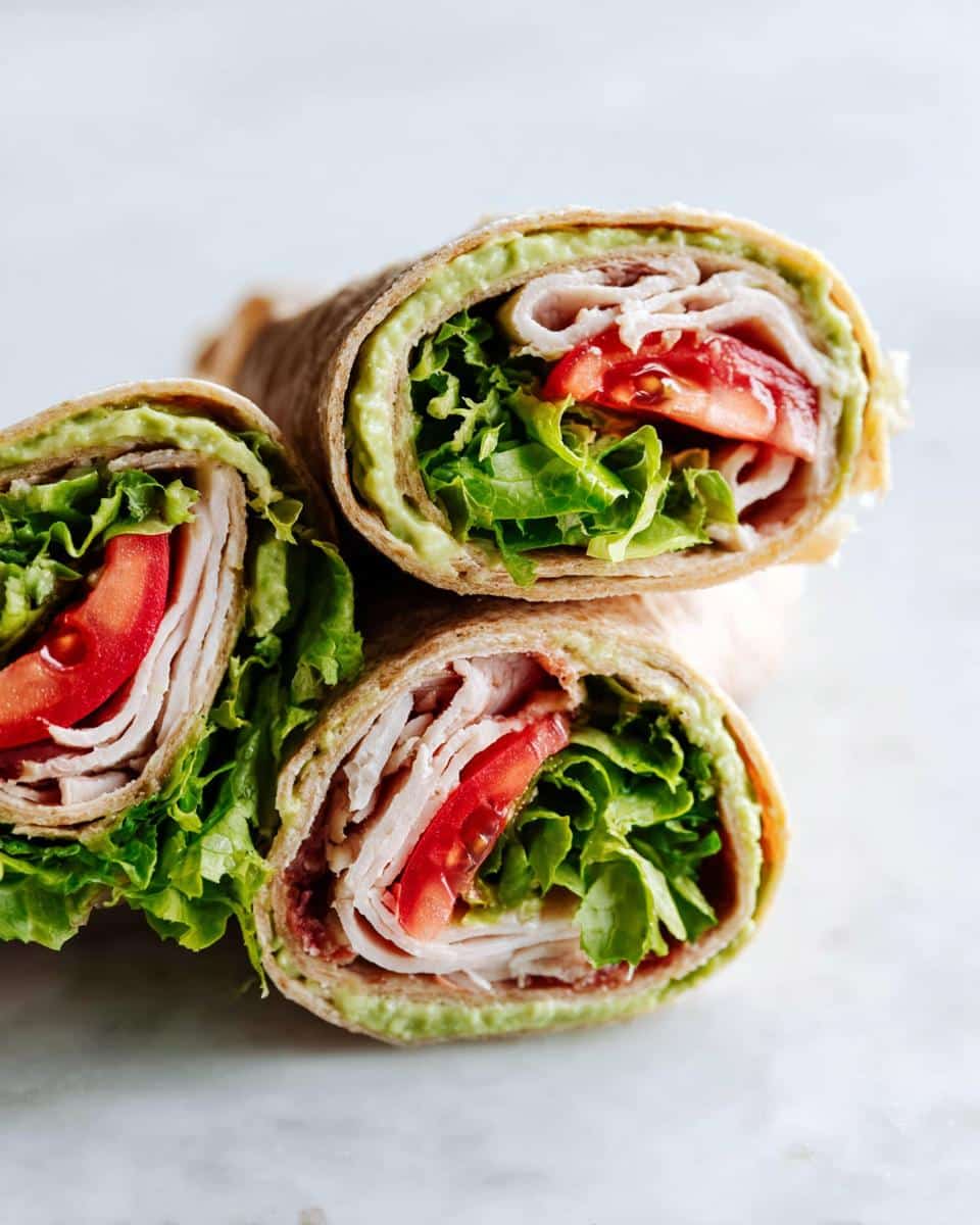 Three halves of a Turkey & Avocado Whole Wheat Wrap stacked, showing layers of turkey, tomato, lettuce, and avocado spread.