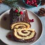 A slice of Vanilla Roll Yule Log with Chocolate Ganache showing the vanilla cake spiral and chocolate filling, garnished with cranberries.