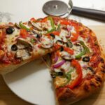 A close-up of a freshly baked Veggie Supreme Pizza with roasted vegetables, showing a cheesy pull as a slice is lifted.