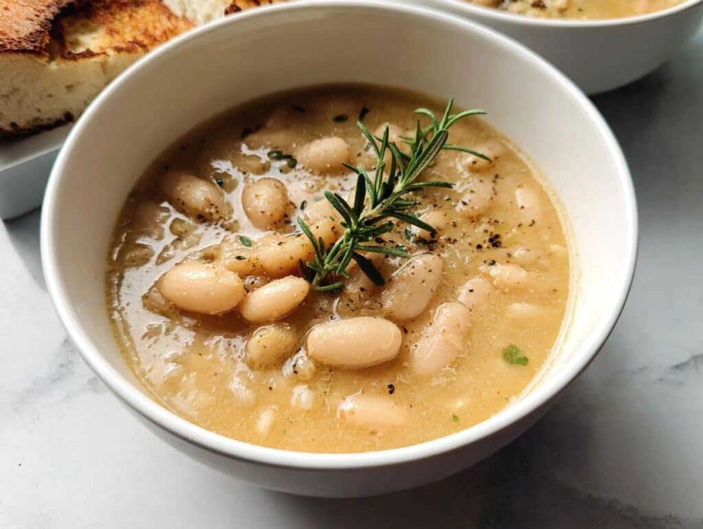 Close-up of a bowl of creamy White Bean & Rosemary Soup garnished with a fresh rosemary sprig and black pepper.