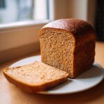 A freshly baked Wholemeal Thermomix Sandwich Loaf sitting on a white plate next to one perfectly cut slice.
