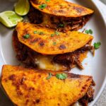 Three crispy, orange-red Authentic Birria Tacos filled with shredded beef and melted cheese, garnished with cilantro and lime wedges.