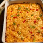 A freshly baked, golden-orange Cajun Crab Dip topped with chopped green onions and red peppers in a white rectangular dish.