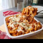 A fork lifting a cheesy, saucy portion of Baked Chicken Ziti Casserole from a white baking dish.