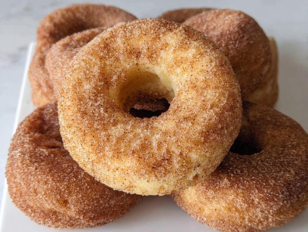 A close-up of several delicious baked cinnamon sugar donuts piled on a white plate.