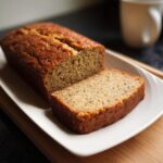 A loaf of moist Banana Bread with No Eggs, with one thick slice cut and resting next to it on a white rectangular plate.