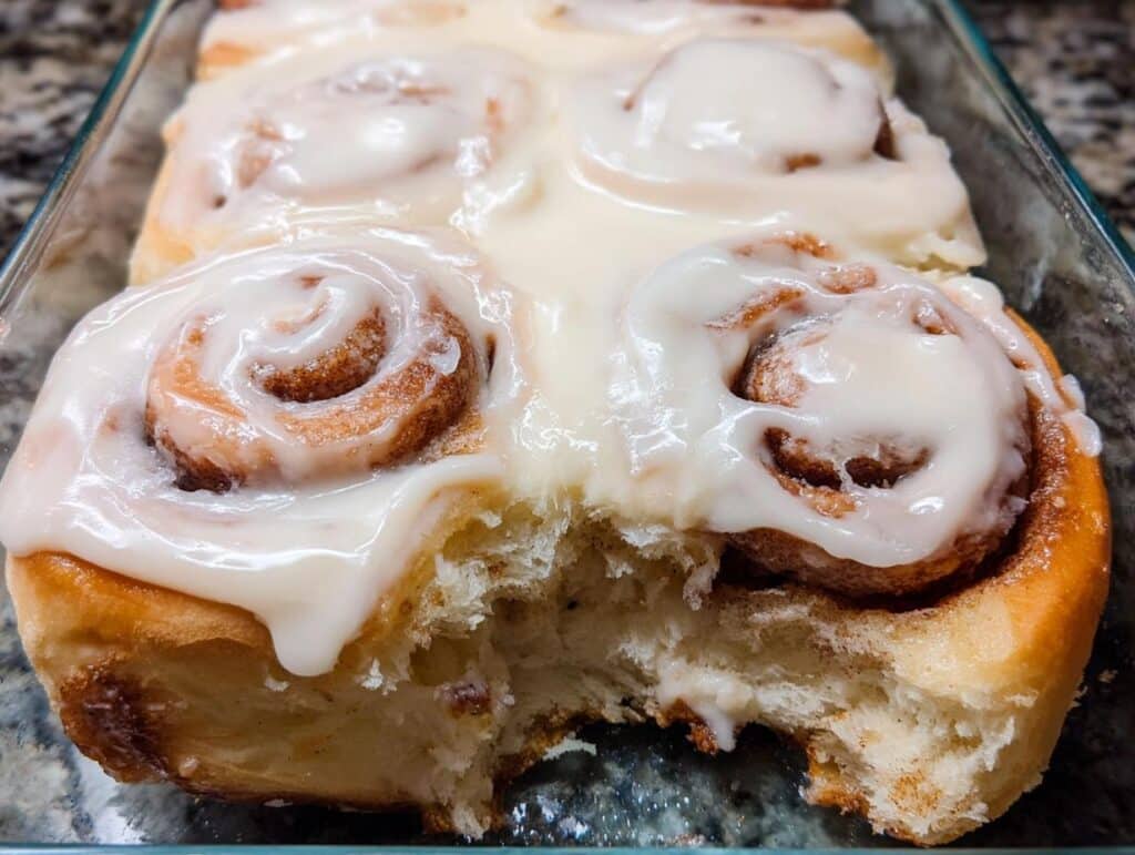 Close-up of gooey, soft homemade cinnamon rolls covered in thick white icing, one roll has a bite taken out.