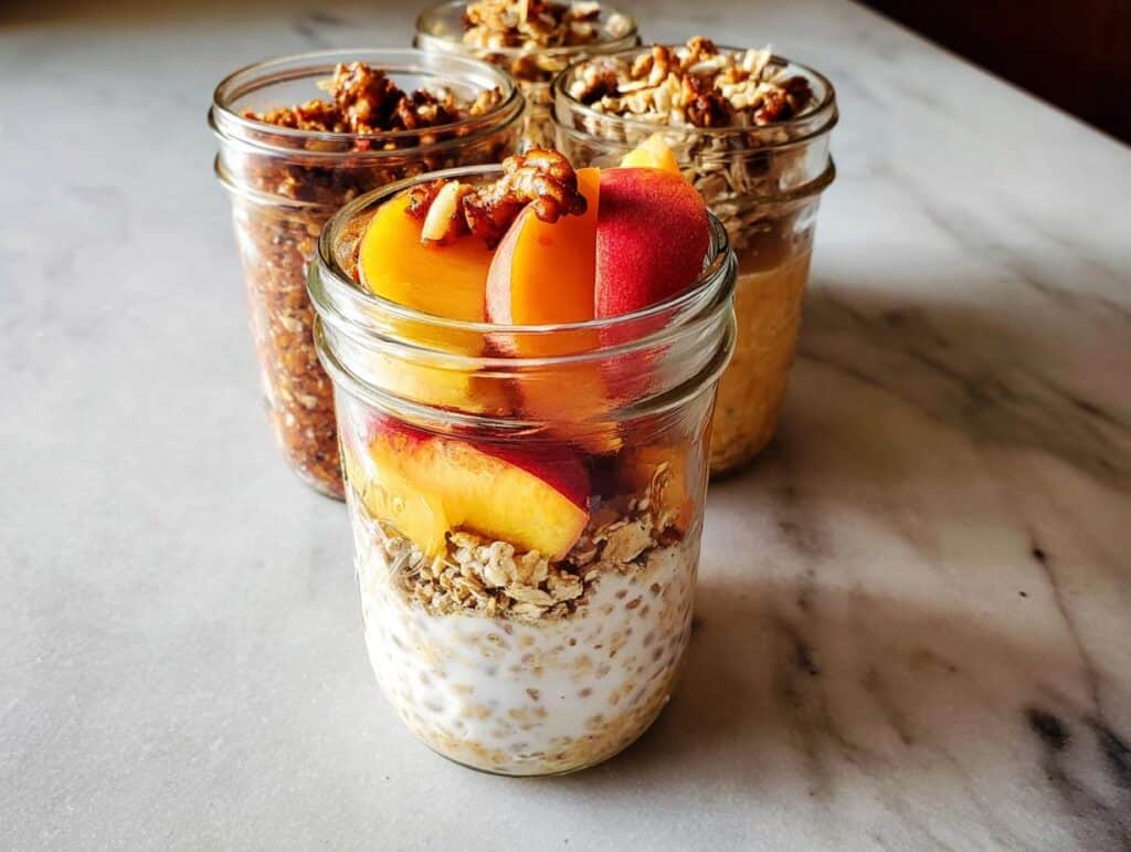 Four jars showing variations of the Best Overnight Oats Recipe, with the front jar topped with fresh peach slices and walnuts.