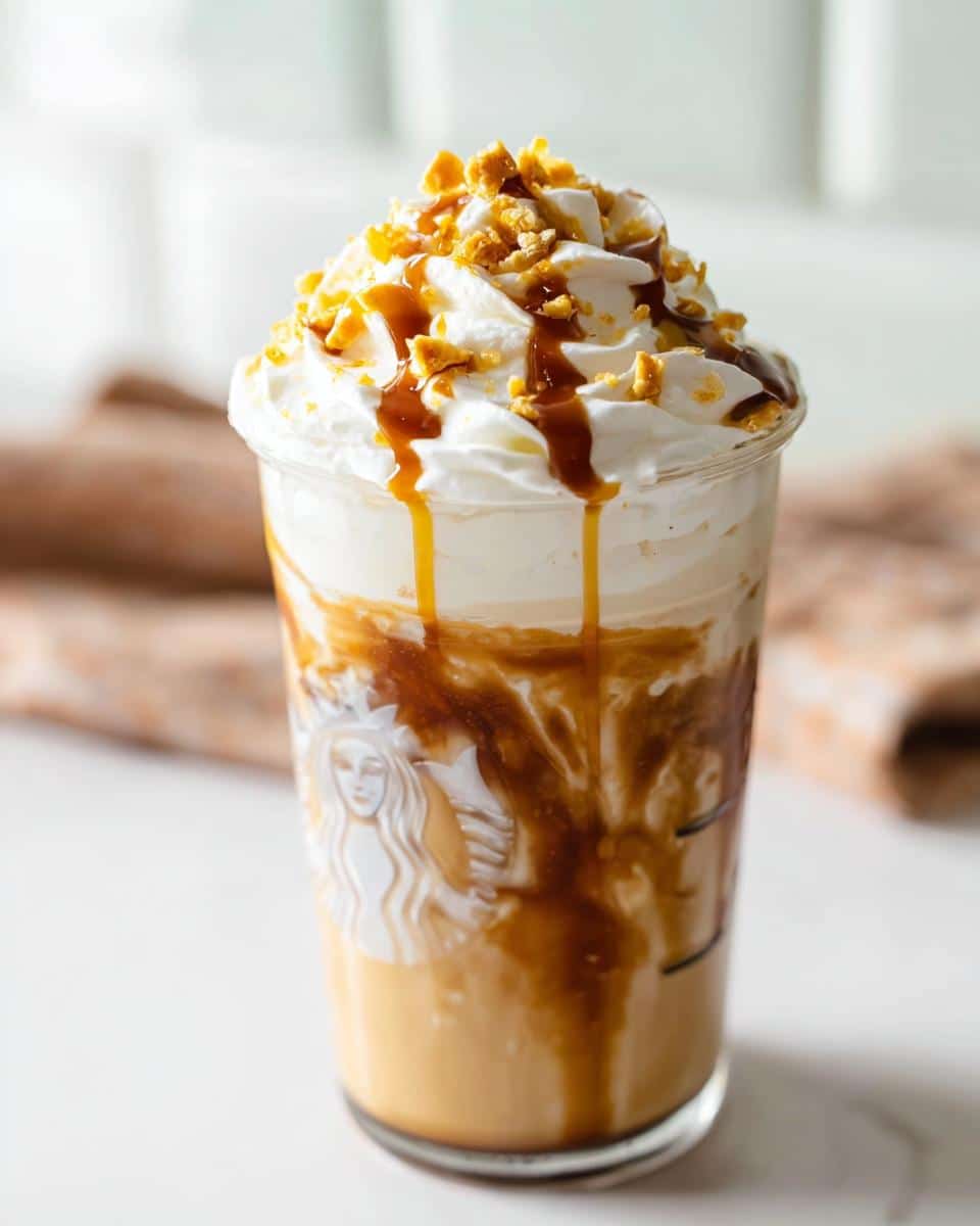 A tall glass of Caramel Ribbon Crunch Frap Copycat topped with whipped cream, caramel drizzle, and crunchy topping.