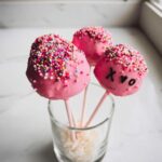 Three pink-dipped Chocolate Valentine Cake Pops decorated with colorful sprinkles, one marked with 'XOXO'.