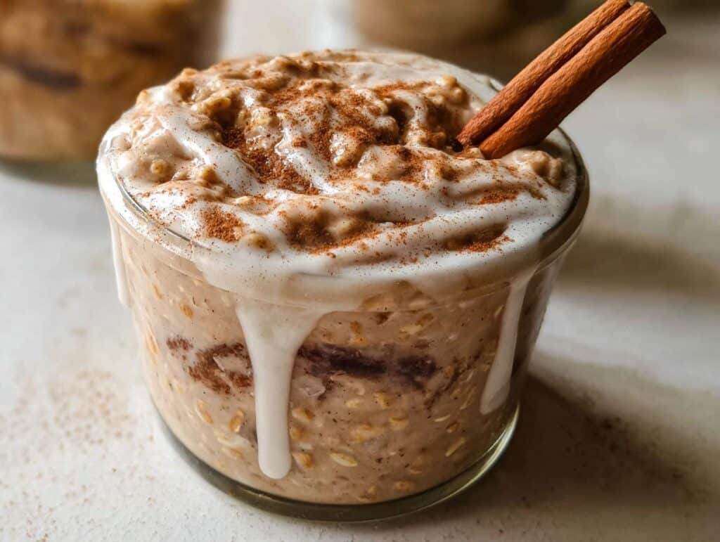 A close-up of Cinnamon Roll Overnight Oats in a glass jar, topped with white glaze, cinnamon powder, and a cinnamon stick.