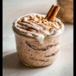 Close-up of Cinnamon Roll Overnight Oats topped with white frosting, nuts, and a cinnamon stick.