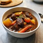 A close-up of a white bowl filled with rich, dark Classic Beef Stew featuring tender beef chunks, carrots, and potatoes, garnished with thyme.