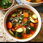 Close-up of a bowl of vibrant Clean Eating Minestrone Soup loaded with carrots, zucchini, and topped with pine nuts.