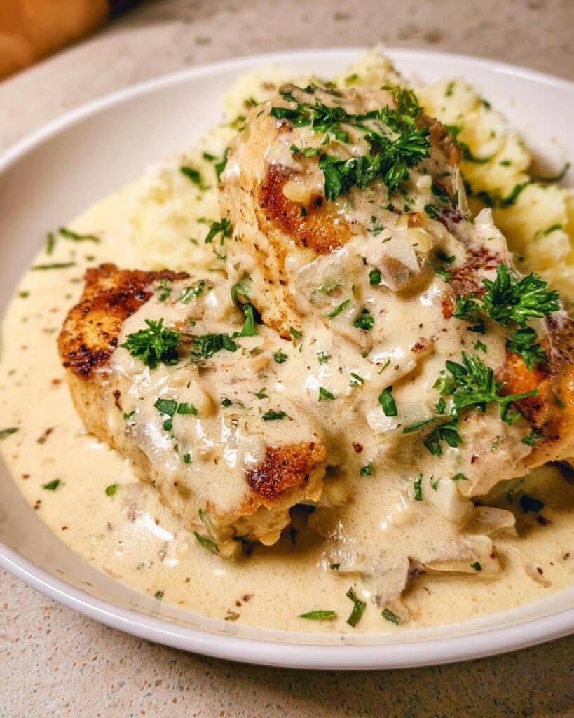 Two golden-brown chicken breasts smothered in creamy garlic sauce and fresh parsley, served with mashed potatoes.