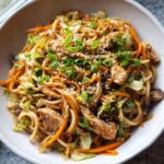 A close-up bowl of Easy Chicken Lo Mein Recipe, featuring noodles, chicken pieces, shredded carrots, cabbage, and topped with sesame seeds and green onions.