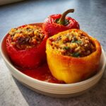 Three cooked Easy Stuffed Bell Peppers with Rice and Beef, one yellow and two red, served in a white bowl with tomato sauce.