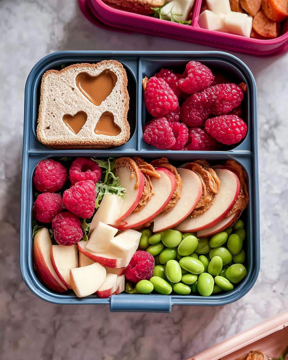 A blue bento box filled with an Easy Valentine Lunch Bento for Kids, featuring heart-shaped sandwich, raspberries, apples with peanut butter, and edamame.
