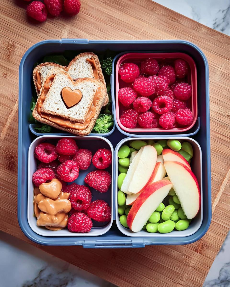Overhead view of an Easy Valentine Lunch Bento for Kids featuring a heart-cutout sandwich, raspberries, edamame, and apple slices.