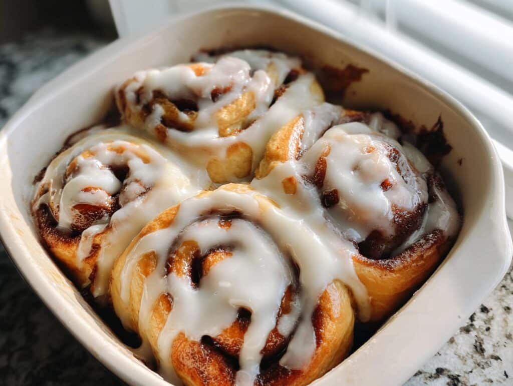 Close-up of four freshly baked, fluffy cinnamon rolls covered in thick white icing in a white baking dish.