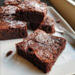 Close-up of fudgy Eggless Chocolate Brownies squares dusted with powdered sugar on a white plate.