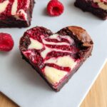 Close-up of a fudgy Heart Shaped Brownies featuring a vibrant red and white cream cheese swirl, garnished with fresh raspberries.