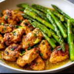 Close-up of golden brown Garlic Butter Chicken Skillet pieces swimming in sauce next to bright green asparagus.